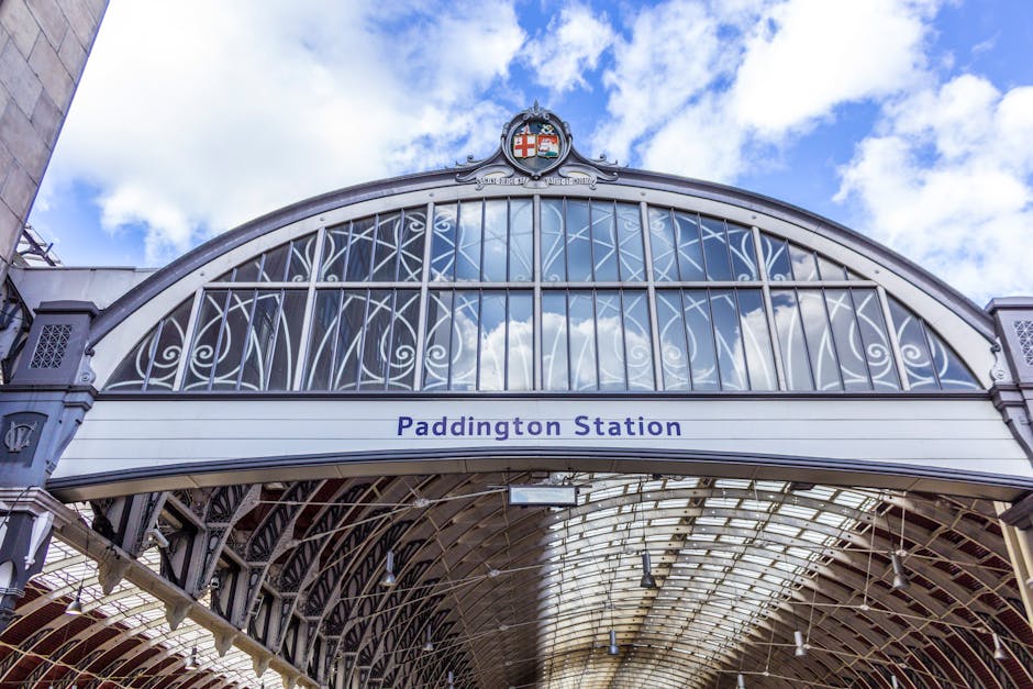 A detailed exterior view of Paddington Station's ornate metal archway and glass roof structure, displaying intricate white metal latticework and decorative crest at the top. The station sign with blue lettering reads 'Paddington Station' beneath the arch. The roof's glass panels reflect a partly cloudy sky with white clouds and blue patches. Inside, the covered platform area is visible, with metal supports and lighting fixtures. This image captures the architectural features of the historic railway station in London, associated with house removals and home relocation services, illustrating the classic setting for furniture transport and moving logistics managed by companies like Man with Van Paddington.
