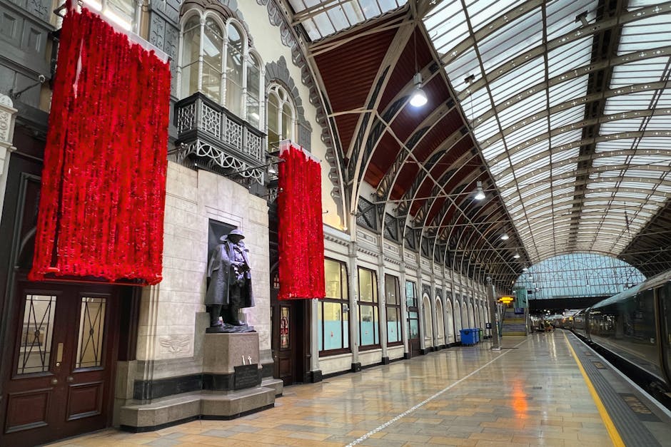 Inside a historic train station with a curved, glass-paneled roof and ornate architectural details, including decorative ironwork and arched windows. The platform is empty, with a row of closed doors along the wall and two large red floral arrangements hanging on either side of a bronze statue of a man in a coat and hat. The polished wooden floor reflects the station’s lighting, and a set of blue recycling bins are positioned near the wall. On the right, a waiting train is visible on the track, with the station's modern glass entrance visible in the distance. The scene captures the environment typical of home relocation and furniture transport processes, with the station as a destination or origin for moving services provided by companies like Man with Van Paddington.