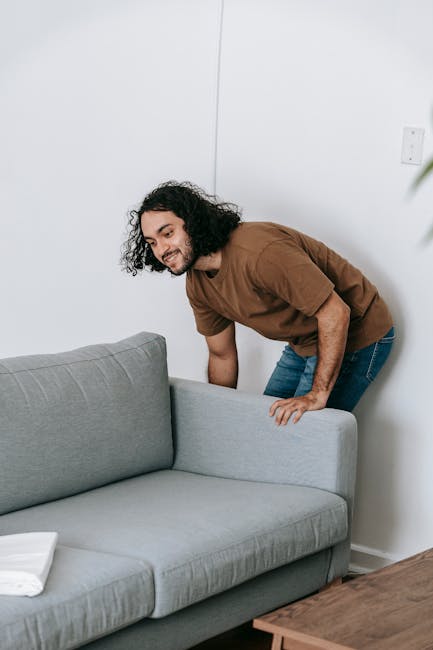 A man with curly dark hair, dressed in a brown t-shirt and blue jeans, is seen carefully lifting a light grey fabric sofa at the edge of a room with white walls. The sofa appears to be part of a home relocation process, and the man is using both hands to support the piece as he prepares to carry or transport it. In the foreground, a small wooden table is partially visible, with a neatly folded white cloth or sheet resting on top. The environment is well-lit, suggesting natural light, and the scene captures a moment of furniture dismantling or loading for removal, typical of professional removals services like those offered by Man with Van Paddington. The overall setting indicates interior space ready for furniture transport, with the focus on efficient handling of bulky items in a moving or packing scenario.