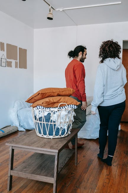 Inside a residential room during a home relocation, two individuals are engaged in packing or unpacking activities as part of furniture and belongings moving process. A man with dark, curly hair tied back, dressed in a red long-sleeve top and dark trousers, stands near a table with a large laundry basket filled with cushions, blankets, and pillows wrapped in various textiles including a checkered towel. A woman with curly hair, wearing a grey hoodie and dark trousers, is turned away from the camera, possibly managing packing materials near a wrapped piece of furniture or large object covered in protective white padding. The room has wooden flooring, white walls, and a ceiling light fixture. On the wall, there are some taped or painted patches, indicating ongoing decor or preparation for moving. Visible on the window-side, the space is illuminated by natural light, suggesting daytime. The scene captures a typical moment in furniture transport, packing, and safe removal as part of house removals offered by Man with Van Paddington.