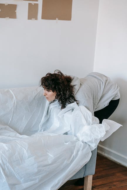 A man with curly dark hair, dressed in a brown t-shirt and blue jeans, is seen carefully lifting a light grey fabric sofa at the edge of a room with white walls. The sofa appears to be part of a home relocation process, and the man is using both hands to support the piece as he prepares to carry or transport it. In the foreground, a small wooden table is partially visible, with a neatly folded white cloth or sheet resting on top. The environment is well-lit, suggesting natural light, and the scene captures a moment of furniture dismantling or loading for removal, typical of professional removals services like those offered by Man with Van Paddington. The overall setting indicates interior space ready for furniture transport, with the focus on efficient handling of bulky items in a moving or packing scenario.