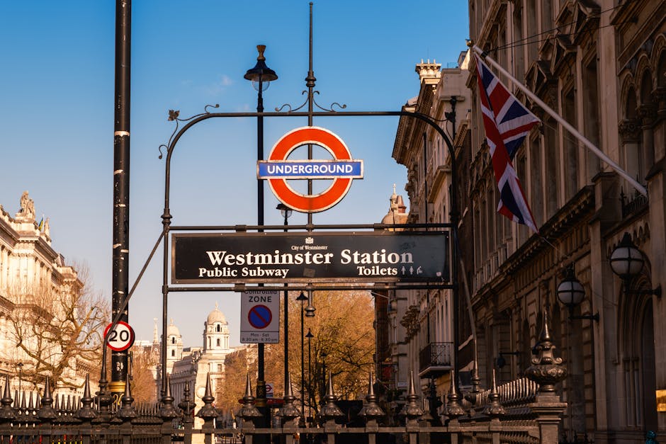 Exterior view of Westminster Station in London with a prominent red and blue London Underground sign hanging above the station entrance. Below the sign, a black rectangular sign displays 'Westminster Station' and indicates available facilities such as public subway access, toilets, and the city of Westminster. To the right, a Union Jack flag is visible, fluttering in the wind, attached to a flagpole mounted on an ornate stone building with architectural details. In the foreground, a metal fence with decorative elements runs along the pavement. Several traffic signs, including a 20 mph speed limit and a no parking zone, are mounted on poles nearby. The sky is clear with bright sunlight illuminating the scene, and trees with autumn-colored leaves are visible behind the signs. The image captures an urban environment typical of London, relevant to the logistics of home relocation or furniture transport, which [COMPANY_NAME], Man with Van Paddington, might assist with when arranging legal parking or moving permits for house removals.