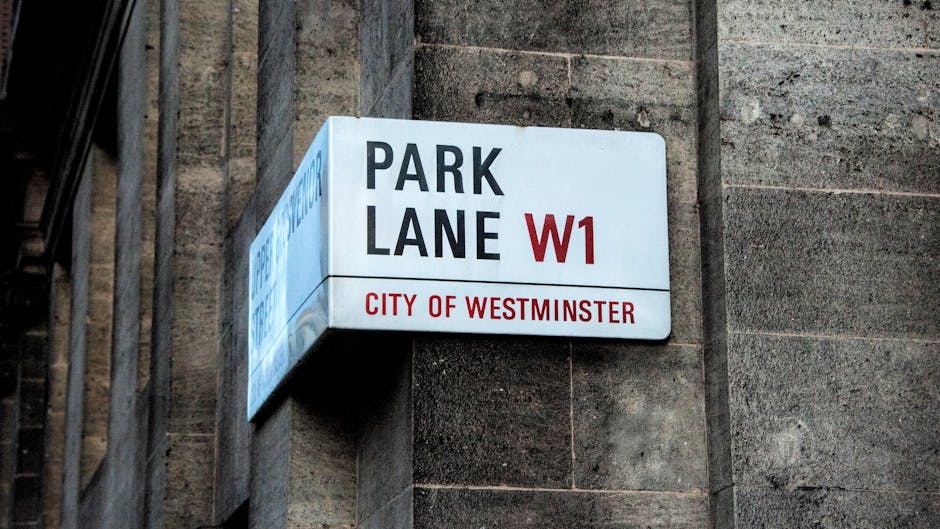 Photograph of a rectangular white and red street sign mounted on a dark brick wall in Westminster, displaying the words 'PARK LANE W1' and 'CITY OF WESTMINSTER'. The sign indicates the previously designated parking lane for the area. The background includes a metal railing on the left side, and the lighting suggests an outdoor urban environment during daytime. This image relates to house removals and moving logistics, as parking permissions and suspensions are often necessary for local home relocation and furniture transport. The presence of the sign highlights the importance of understanding parking regulations, such as obtaining a parking suspension, which [COMPANY_NAME] may assist with during full-service removals in the Paddington area, especially when navigating busy city streets to facilitate efficient loading and unloading procedures.