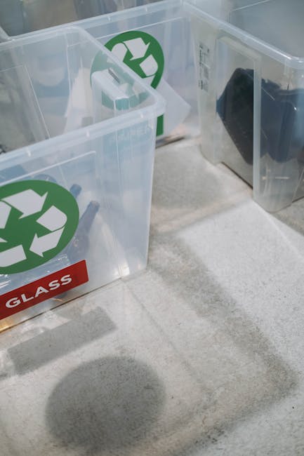 The image displays two clear, rectangular recycling bins made of plastic, positioned on a light-colored carpeted floor indoors, with one bin slightly behind the other. Each bin features a prominent green circular recycling symbol on the front, indicating they are designated for recyclable materials. The bin on the left has a red label with the word 'GLASS' in white capital letters adhered to its front, signifying its use for glass waste, while the bin on the right contains a black item, possibly a pad or bag, visible through the transparent sides. The interior environment is well-lit, and the bins are situated near an unobstructed doorway or opening, suggesting they are placed conveniently for waste disposal or collection. These recycling bins are commonly used during home relocation or packing and moving processes, as part of eco-friendly waste management practices, for companies like Man with Van Paddington providing professional removals and recycling services.