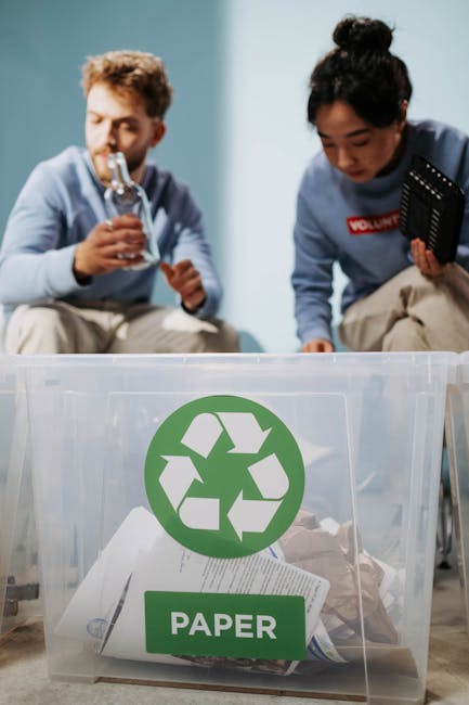 A series of large cardboard boxes, some flattened and others partially assembled, are stacked on a pavement outside a property undergoing a home relocation. The boxes contain various packing materials such as crumpled brown paper, bubble wrap, and plastic wrapping. They are positioned in front of a white metal fence with a prominent blue sign reading ‘RECYCLING ONLY’ featuring a green recycling symbol. Some boxes are stored in brown and blue recycling bins, indicating preparation for eco-friendly waste disposal or donation, part of the packing and moving process managed by Man with Van Paddington. Above the bins, a metal structure possibly part of scaffolding or building framework is visible. The environment is clear and well-lit, with natural daylight illuminating the scene, illustrating typical activity involved in furniture transport, packing, or moving logistics during a house removal.
