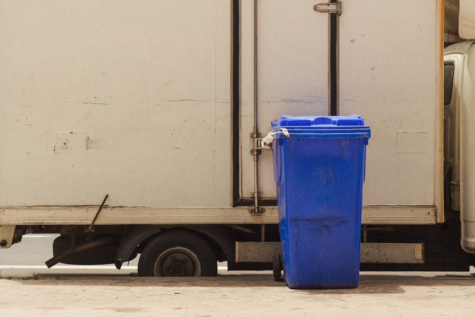 A white commercial van used for house removals, parked on a street with the side door closed. A large blue recycling bin with a closed lid is positioned on the pavement next to the van, partially blocking the view of the vehicle's lower section. The van appears weathered, with visible dirt and minor scratches on its surface. Inside, visible through the open side door, are stacking cardboard boxes, some wrapped in plastic or fabric, and packing materials such as plastic wrap and cardboard protectors. A furniture trolley or dolly is seen near the entrance, used for transporting household items during packing and moving processes. The environment is outdoors with natural lighting, and the setting suggests a home relocation scene, where furniture and boxes are prepared for loading into the van for transportation. Man with Van Paddington’s removal service could be involved in safely moving these household goods, complying with regulations such as those related to waste disposal and permits for unloading.