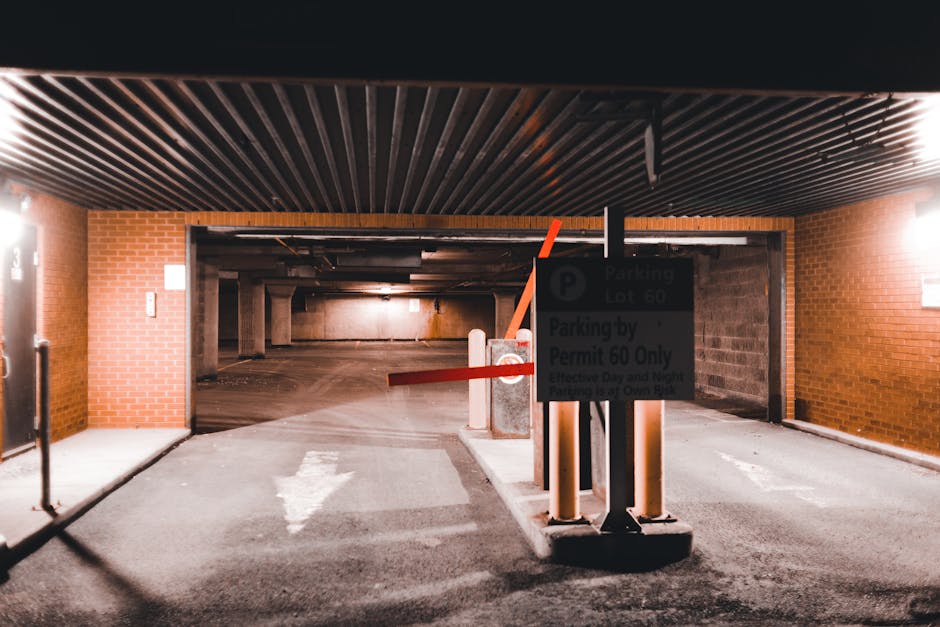 The image shows an underground parking garage entrance with a closed barrier gate and a parking sign indicating parking lot 60 and permit requirements. The entrance is framed by brick walls on either side and a black corrugated ceiling overhead. The barrier arm, painted red and white, is lowered across the vehicle entry point, with a small concrete base supporting it. A white arrow painted on the asphalt directs vehicles into the parking area. Inside the garage, visible concrete pillars and a dimly lit interior extend into the distance, creating a sense of depth. The environment appears clean and well-maintained, suitable for vehicle access and home relocation purposes. This setting could be part of a property in Paddington where [COMPANY_NAME] manages furniture transport and packing during house moves, highlighting the logistics and spatial considerations involved in the removal process, especially concerning compliance with local parking and permit regulations in Paddington.