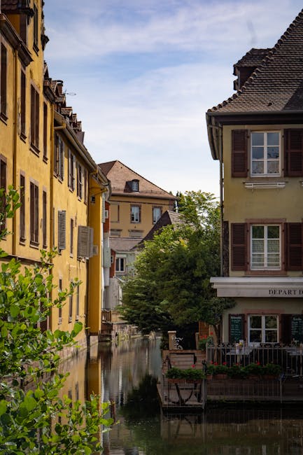 The image shows a narrow residential street in Little Venice, Paddington, featuring traditional European-style buildings with pastel yellow and beige facades, some with wooden shutters. A small canal runs alongside the buildings, reflecting their facades and the partly cloudy blue sky above. In the foreground, green foliage from small trees and shrubs partially frames the scene. Visible in the image are some wrapped furniture pieces and cardboard boxes placed near the buildings, suggesting preparations for home relocation or moving services. The setting is well-lit with natural daylight, highlighting the architectural details and the tranquil canal environment. The professional removal process by Man with Van Paddington is implied through the presence of packing materials and furniture, emphasizing careful packing and loading activities within this picturesque setting.
