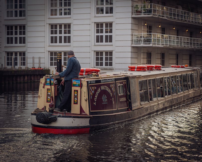 A canal houseboat operated by London Waterbus Company is navigating a narrow waterway in Paddington, with a male crew member standing at the stern managing the vessel's controls during daylight hours. The boat is painted in cream and maroon colors with decorative floral accents, and has red lifebuoys secured on the roof, which also holds various equipment and safety gear. Behind the boat, a modern white residential building with multiple windows and balconies lines the canal, indicating an urban environment with accessible waterway routes suited for moving and furniture transport during a home relocation. The water reflects the boat and building, emphasizing the confined, picturesque setting typical of Paddington’s canal network, which often requires careful navigation and strategic planning for removals and moving services by companies such as Man with Van Paddington.