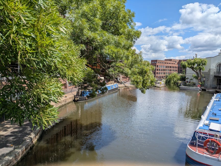 The image shows a narrow residential street in Little Venice, Paddington, featuring traditional European-style buildings with pastel yellow and beige facades, some with wooden shutters. A small canal runs alongside the buildings, reflecting their facades and the partly cloudy blue sky above. In the foreground, green foliage from small trees and shrubs partially frames the scene. Visible in the image are some wrapped furniture pieces and cardboard boxes placed near the buildings, suggesting preparations for home relocation or moving services. The setting is well-lit with natural daylight, highlighting the architectural details and the tranquil canal environment. The professional removal process by Man with Van Paddington is implied through the presence of packing materials and furniture, emphasizing careful packing and loading activities within this picturesque setting.