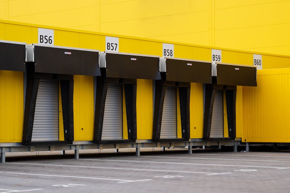 A row of four black and white goods lifts with electronic roll-up doors, numbered B56, B57, B58, and B59, is attached to a bright yellow building exterior. The lifts are positioned at ground level, with their doors closed, and are supported by metal brackets attached to the building's yellow wall. The pavement in front of the loading area is marked with white lines, indicating designated parking or loading zones. Above the lifts, the building's yellow facade extends horizontally, with the numbers displayed on white labels. The lighting appears natural, suggesting daytime conditions. This scene, part of a commercial or industrial building, is related to logistics, packing, and moving operations, consistent with house removals and furniture transport services as offered by Man with Van Paddington.