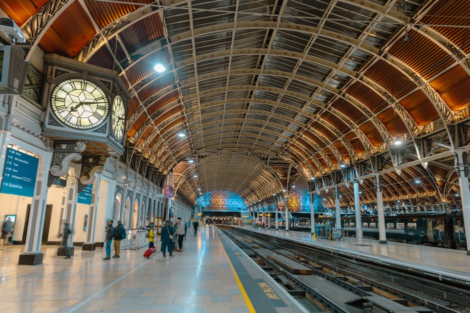 Inside an ornate, spacious train station with a high arched ceiling made of metal and glass, illuminated by several ceiling lights and decorative lighting features. On the left side, vintage-style wall clocks are mounted on the white panelled walls, alongside blue directional signs indicating platform information. Several passengers are present, some carrying luggage or pulling suitcases, dressed in casual and business attire. The platform is clean with a tactile yellow safety line along the edge where train tracks run parallel, ready for the loading process. In the background, train carriages are stationed at the platform, with some equipment such as trolleys visible nearby. The environment appears well-lit and busy, reflecting a typical setting for home relocation or furniture transport arrangements at a major railway station, supporting the movement of household items for house removals or packing and moving services, as managed by professional removal companies like Man with Van Paddington.