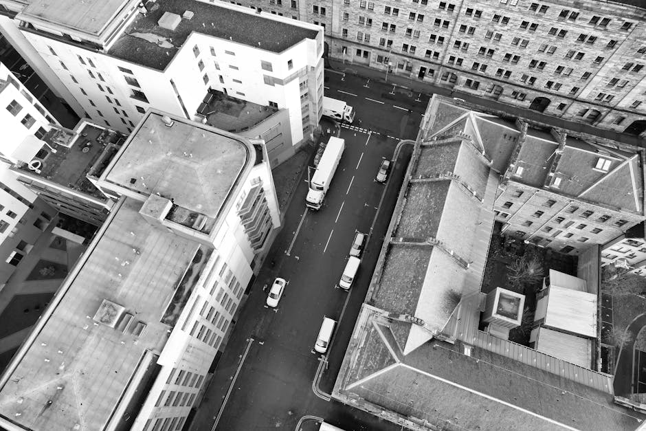 An aerial black-and-white photograph shows a city street lined with multi-storey buildings, with vehicles including cars and vans parked along the curb and moving along the road. At the top, there's a clear view of rooftops with various features such as vents, skylights, and flat surfaces. The street appears to be in a densely built urban area, with narrow sidewalks and closely spaced structures. In the middle of the image, a white van is positioned near the entrance to a building, suggesting a furniture transport or home relocation process. Nearby, several stacked cardboard boxes and wrapped furniture items are visible, indicating packing and loading activities associated with house removals. A trolley or dolly can be seen near the boxes, supporting furniture transport efforts. The image depicts the loading process for a same-day emergency removals service in Paddington, with Man with Van Paddington potentially engaged in moving household items from the building to the vehicle parked on the street, emphasizing efficient planning and logistics in urban house removals.